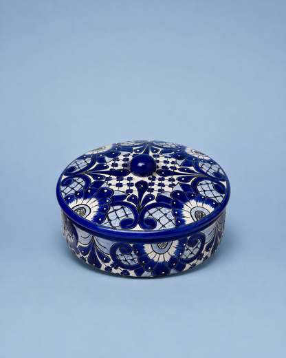A round lidded ceramic box with intricate cobalt blue and white floral patterns, photographed on a solid light blue background.