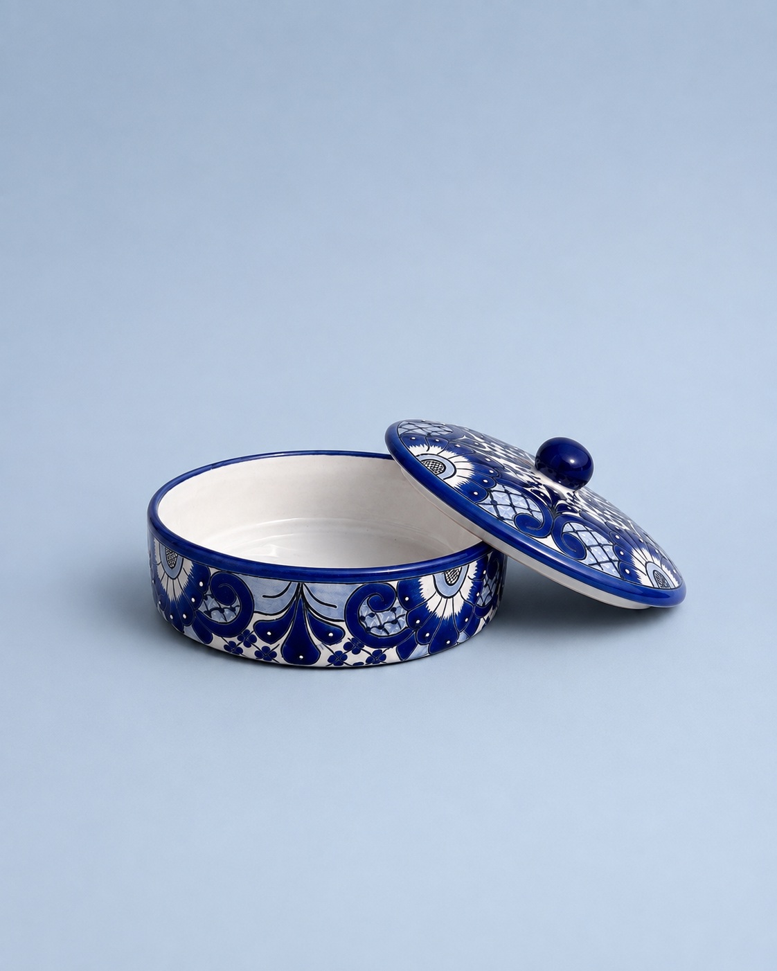 A round ceramic lidded box with a blue and white floral pattern, shown open with the lid propped against it on a light blue background.