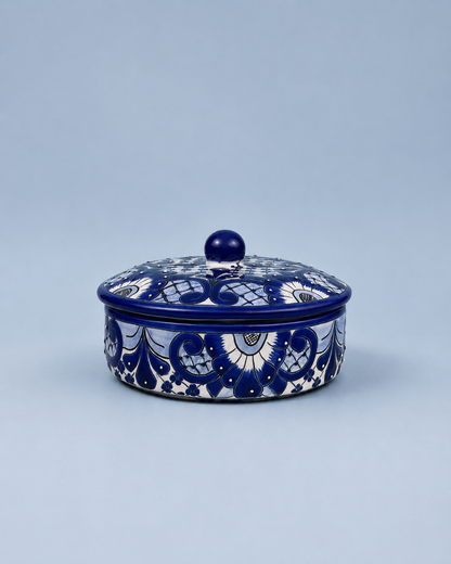 A round lidded ceramic box with a deep cobalt blue and white hand-painted floral pattern, set against a solid light blue-gray background.