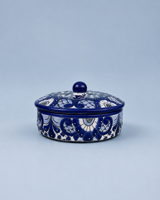 A round lidded ceramic box with a deep cobalt blue and white hand-painted floral pattern, set against a solid light blue-gray background.