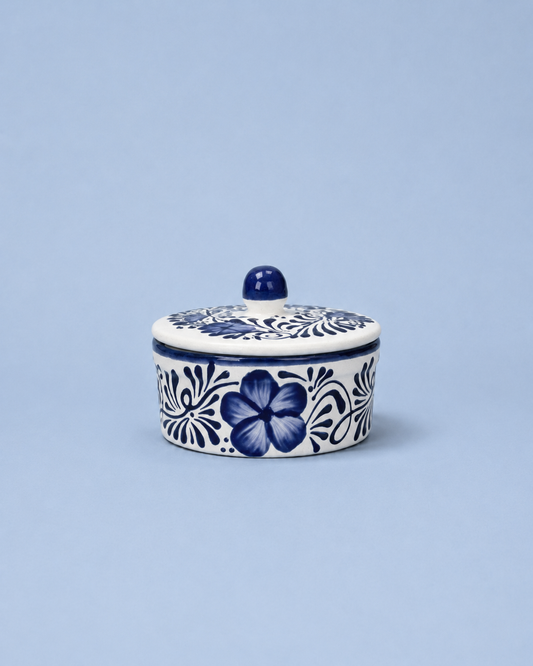 A small round lidded ceramic container with a blue floral pattern on a white background, set against a solid light blue backdrop.