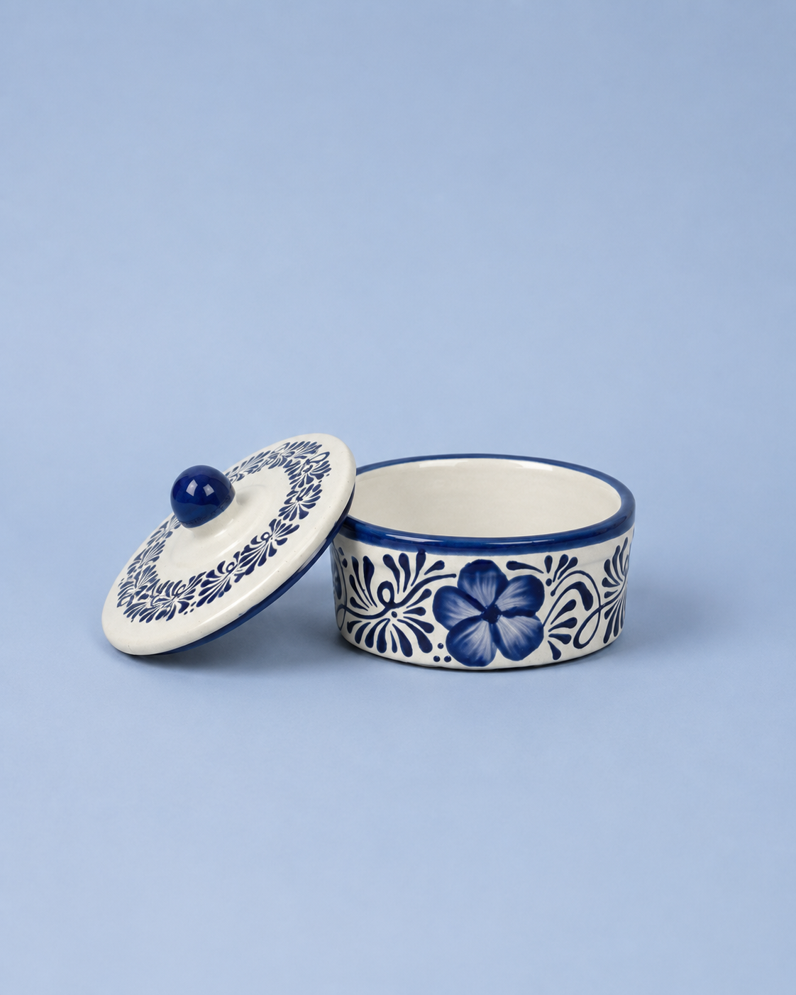 A small round ceramic lidded container with a floral blue and white pattern, shown open with the lid leaning against it on a light blue background.