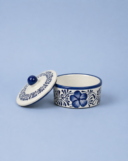 A small round ceramic lidded container with a floral blue and white pattern, shown open with the lid leaning against it on a light blue background.