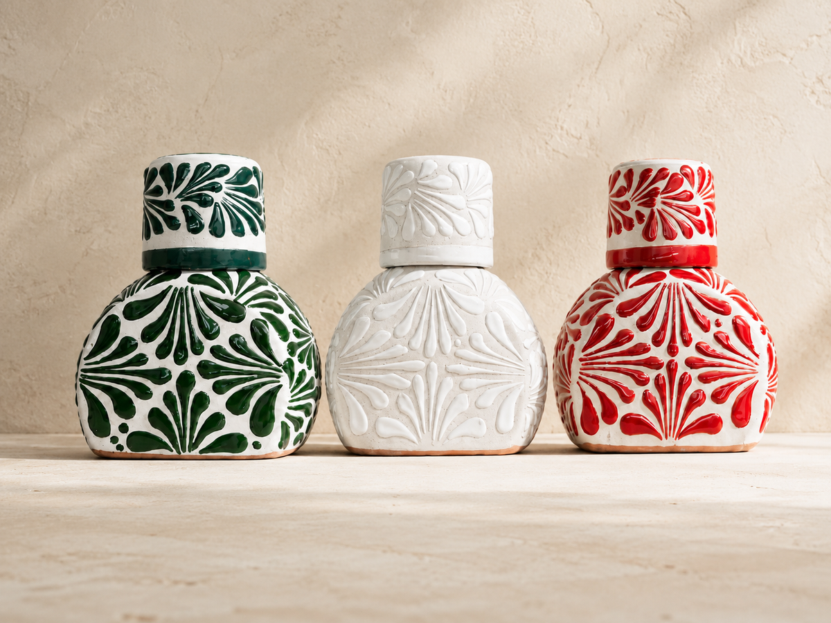 Three ceramic tequila bottles, a green, white, and a red one, with decorative patterns on a clay surface and beige background.