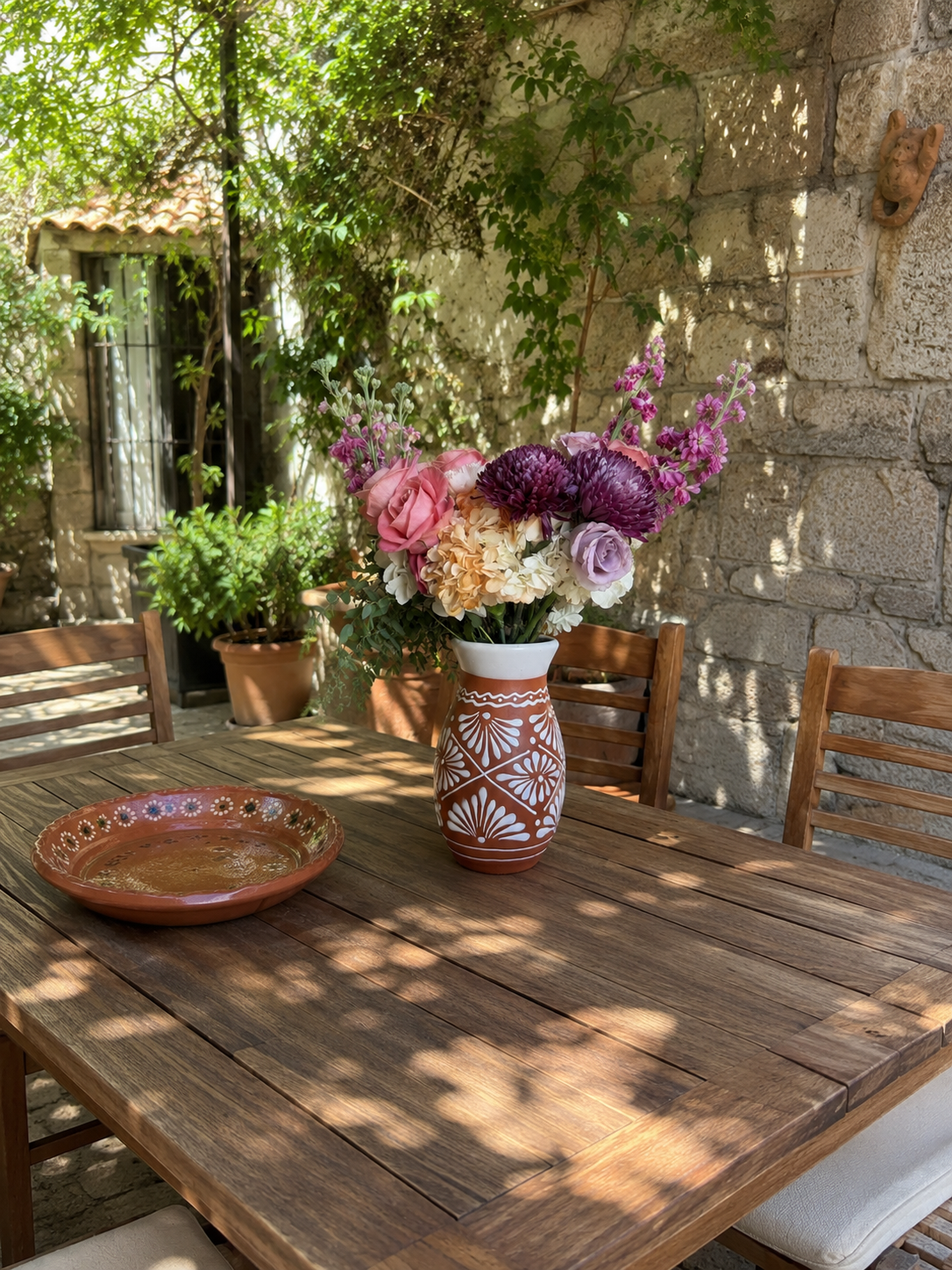 Wooden table with a vase of flowers and a plate outdoors against a stone wall.