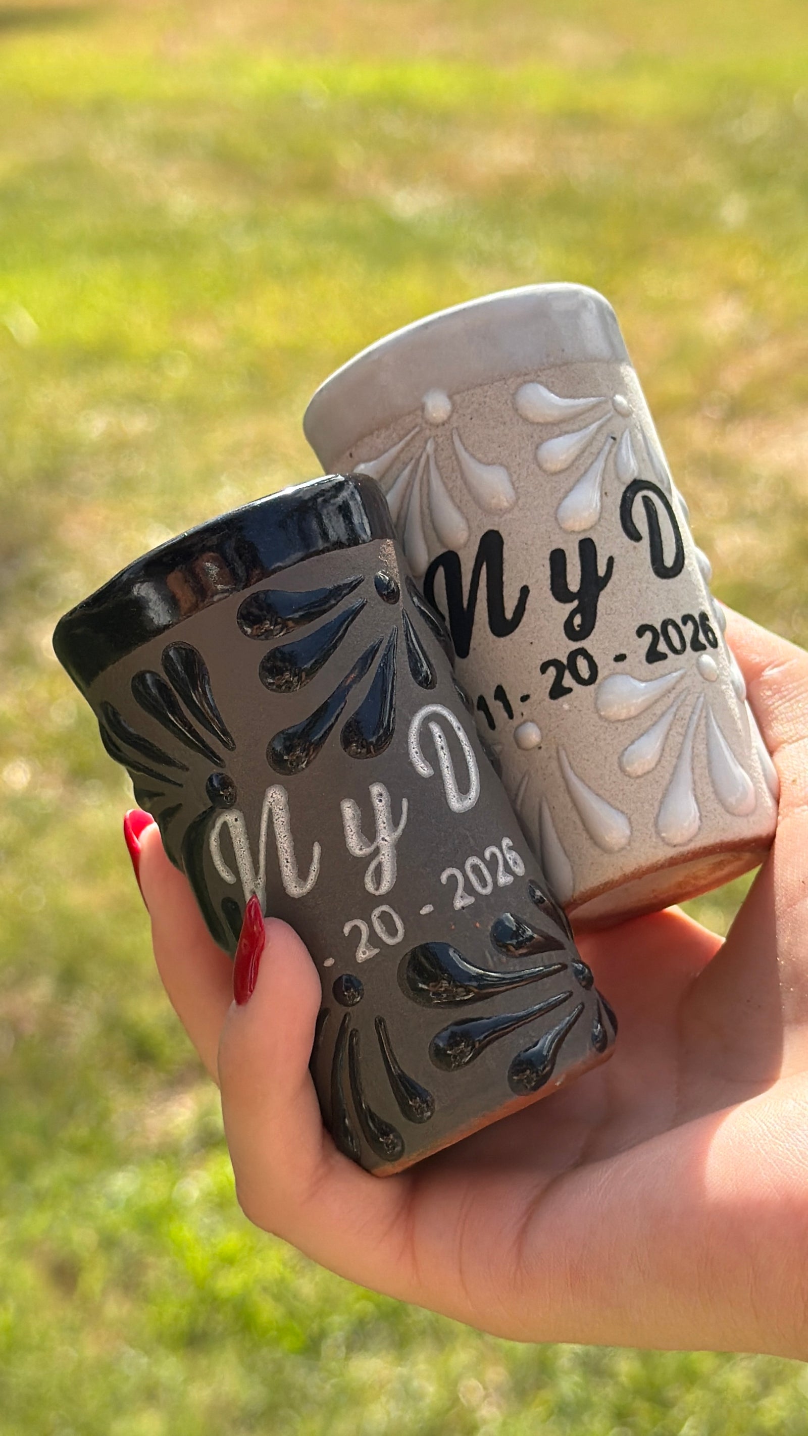 A hand holding two small engraved shot glasses outdoors on a grassy lawn. One shot is dark gray with black floral detailing and the other is light gray with white floral detailing.