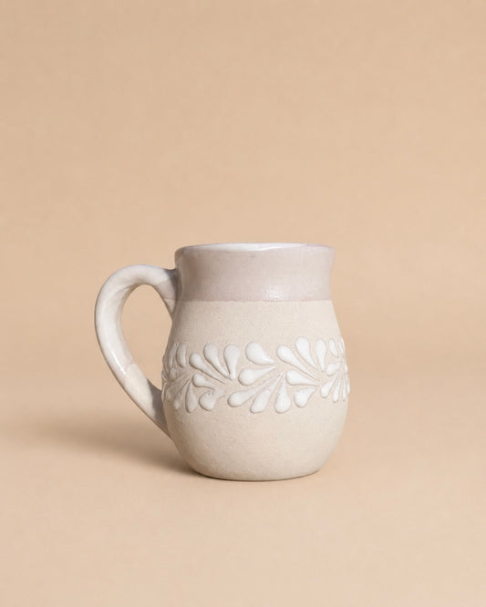 A small ceramic mug with a handle, featuring a matte off-white finish and a raised leaf pattern around the middle, set against a solid beige background.