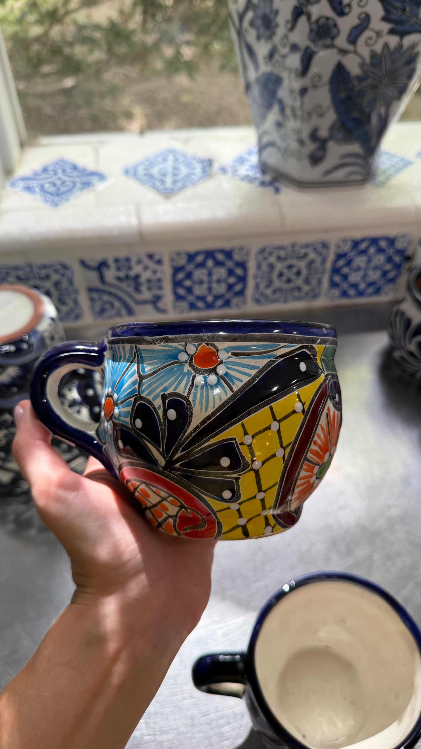 A hand holds a colorful hand-painted ceramic mug with a butterfly design, set against a tiled kitchen background with other ceramic items.